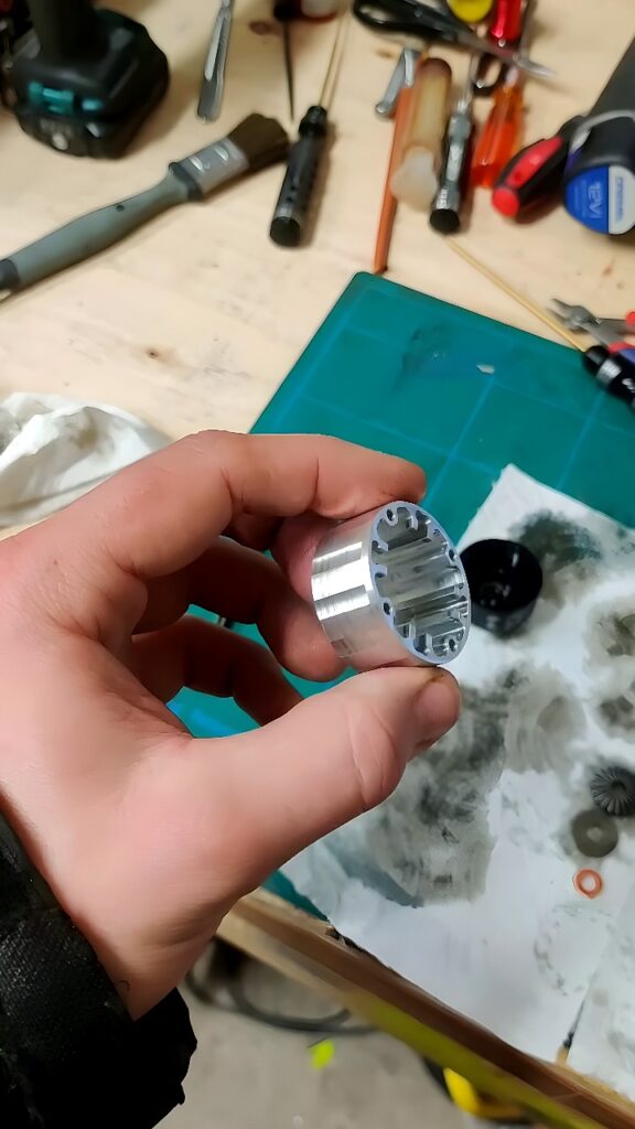 Hand holding a CNC-milled aluminum cylinder with internal slots and bores in a workshop setting