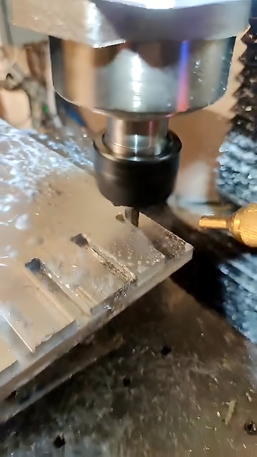 CNC milling machine cutting precise rectangular grooves into a clear plastic sheet with visible chips and coolant nozzle