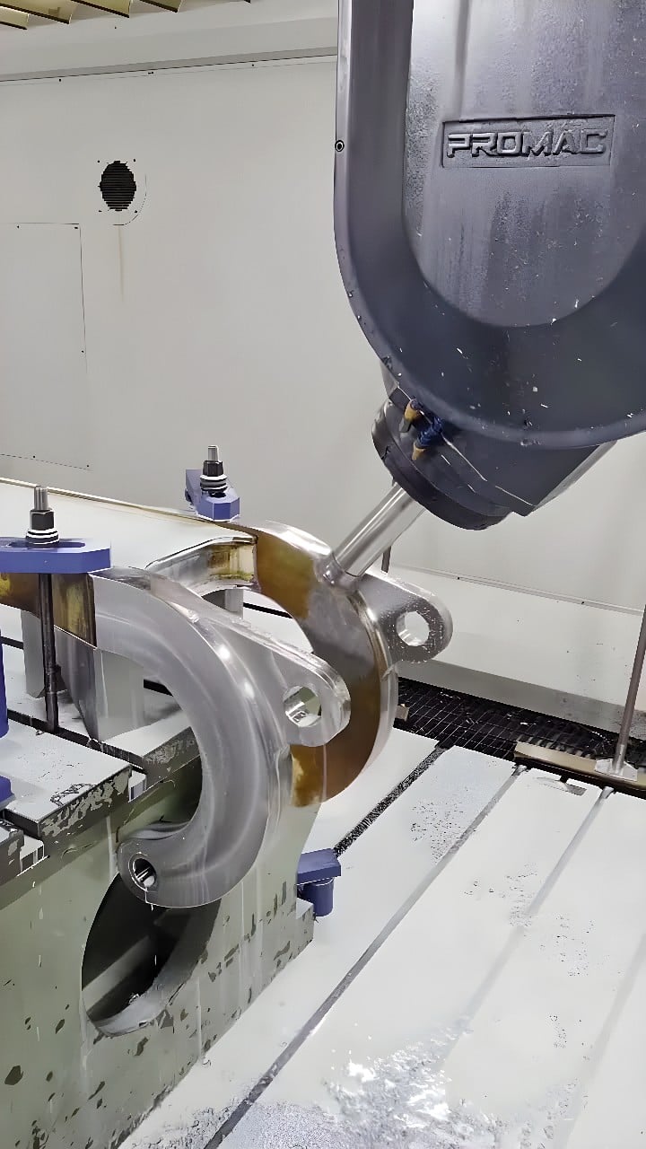 PROMAC 5-axis CNC milling head machining a large aluminum clevis-style bracket clamped in a precision vise, with coolant and metal chips visible