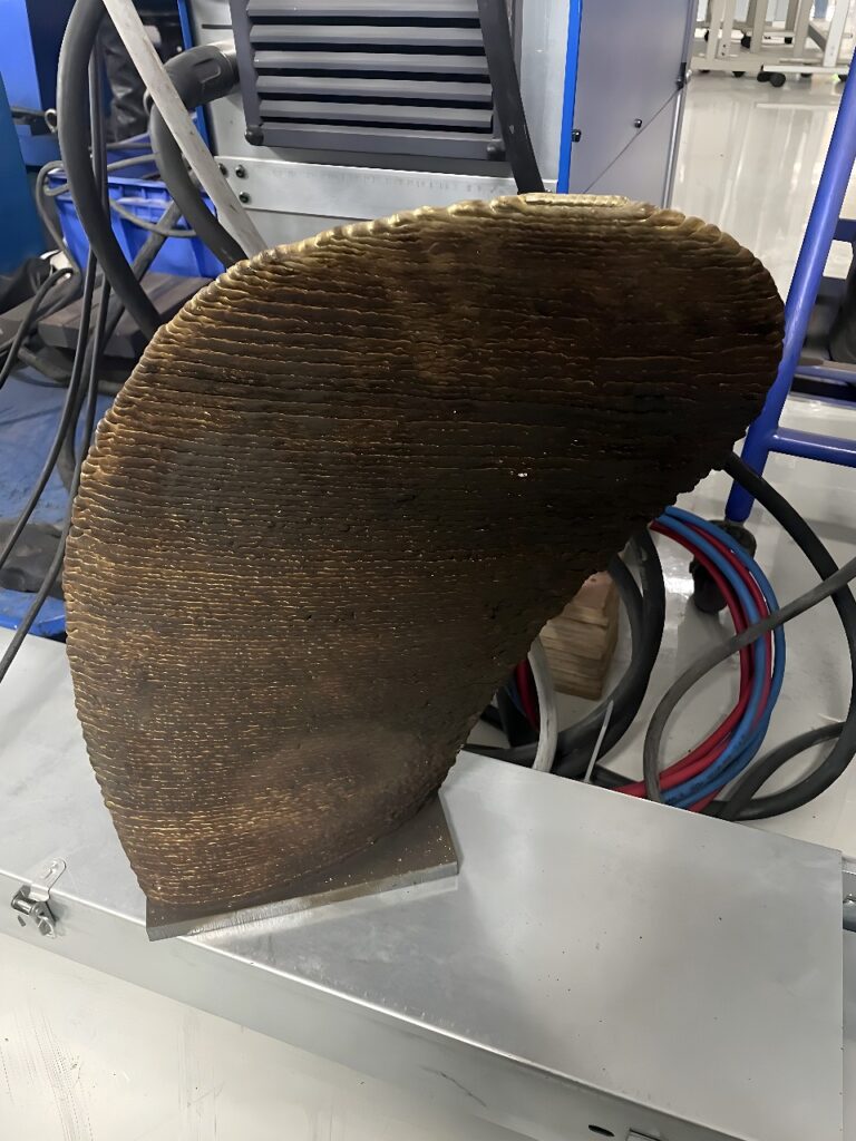 Curved metal workpiece with horizontal layer lines on a large industrial 3D printer build plate, with hoses and cables feeding into the machine