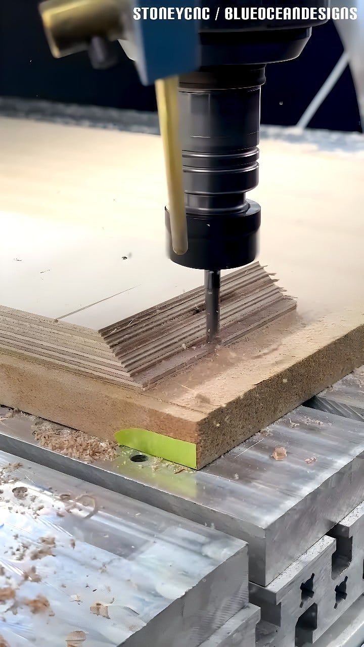 CNC router spindle with straight-flute endmill cutting stepped contours into a stacked plywood panel clamped on an aluminum T-slot table, with wood chips visible.