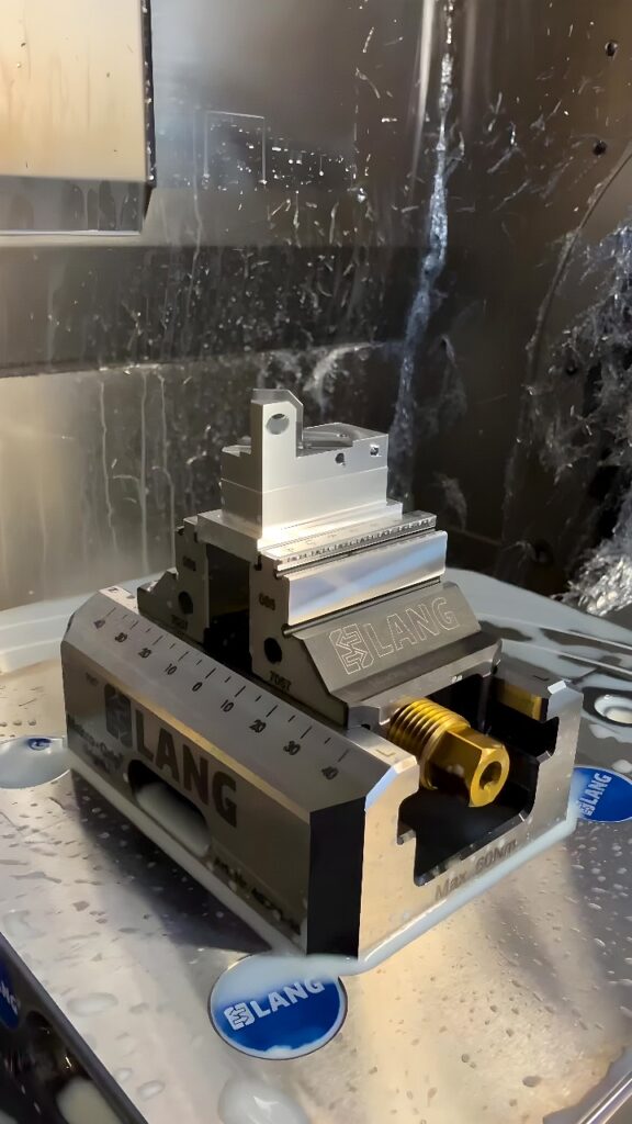 CNC milling center showing a LANG Makro‑Grip vise clamping an aluminum bracket being milled under flood coolant