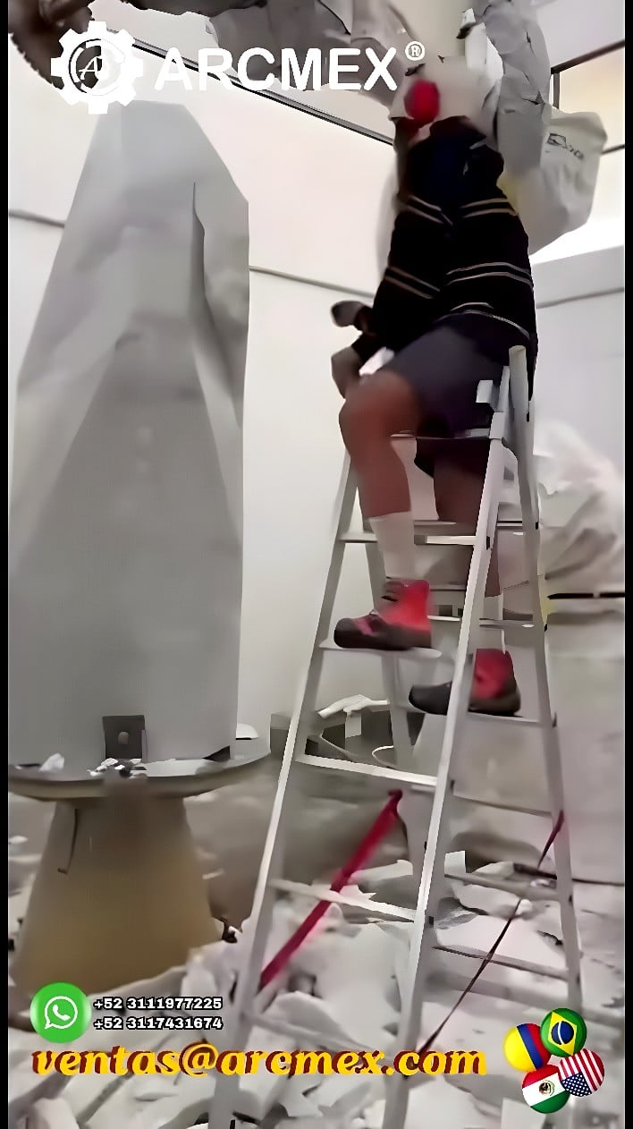 Six-axis robotic arm machining a tall stone obelisk on a powered rotary turntable inside a dust-controlled enclosure with a technician observing