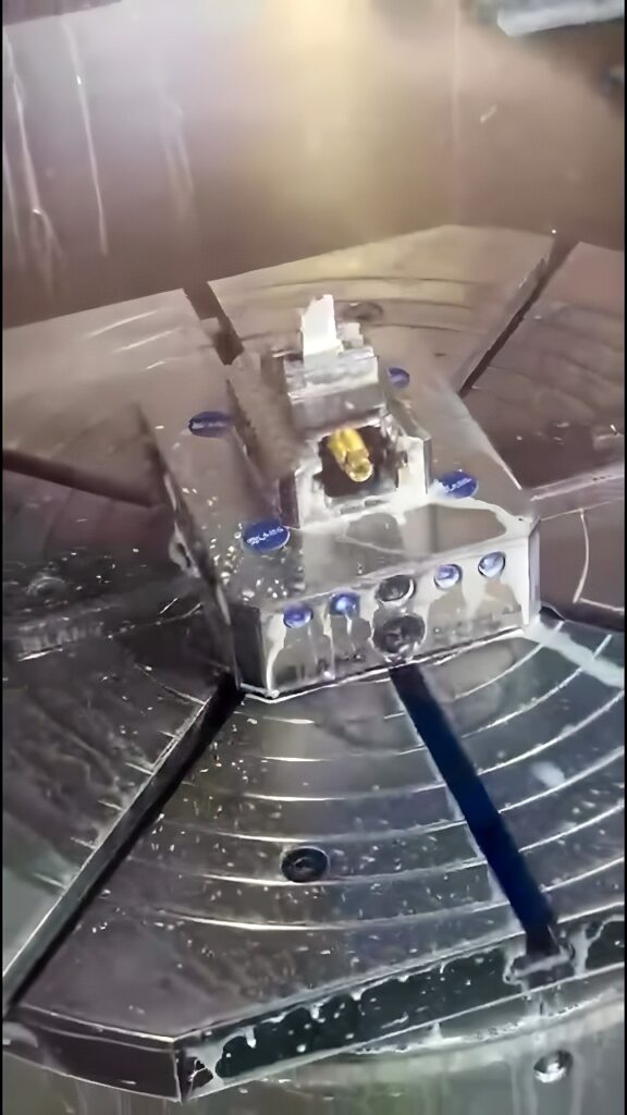 Five-axis CNC machine carving internal pockets and holes into an aluminum housing using a rotary table and coolant-fed end mill