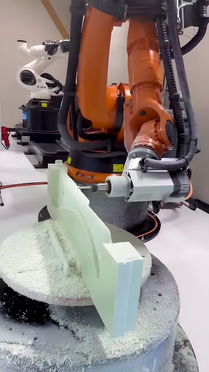 Orange KUKA six-axis robot milling a light green foam block on a rotary table with foam chips collecting below