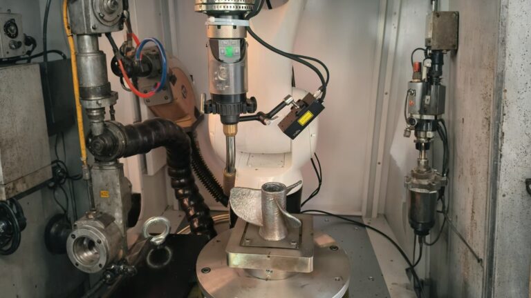 A six-axis industrial robot machining a metal component on a rotary table, with a sensor nearby, in an automated manufacturing cell.